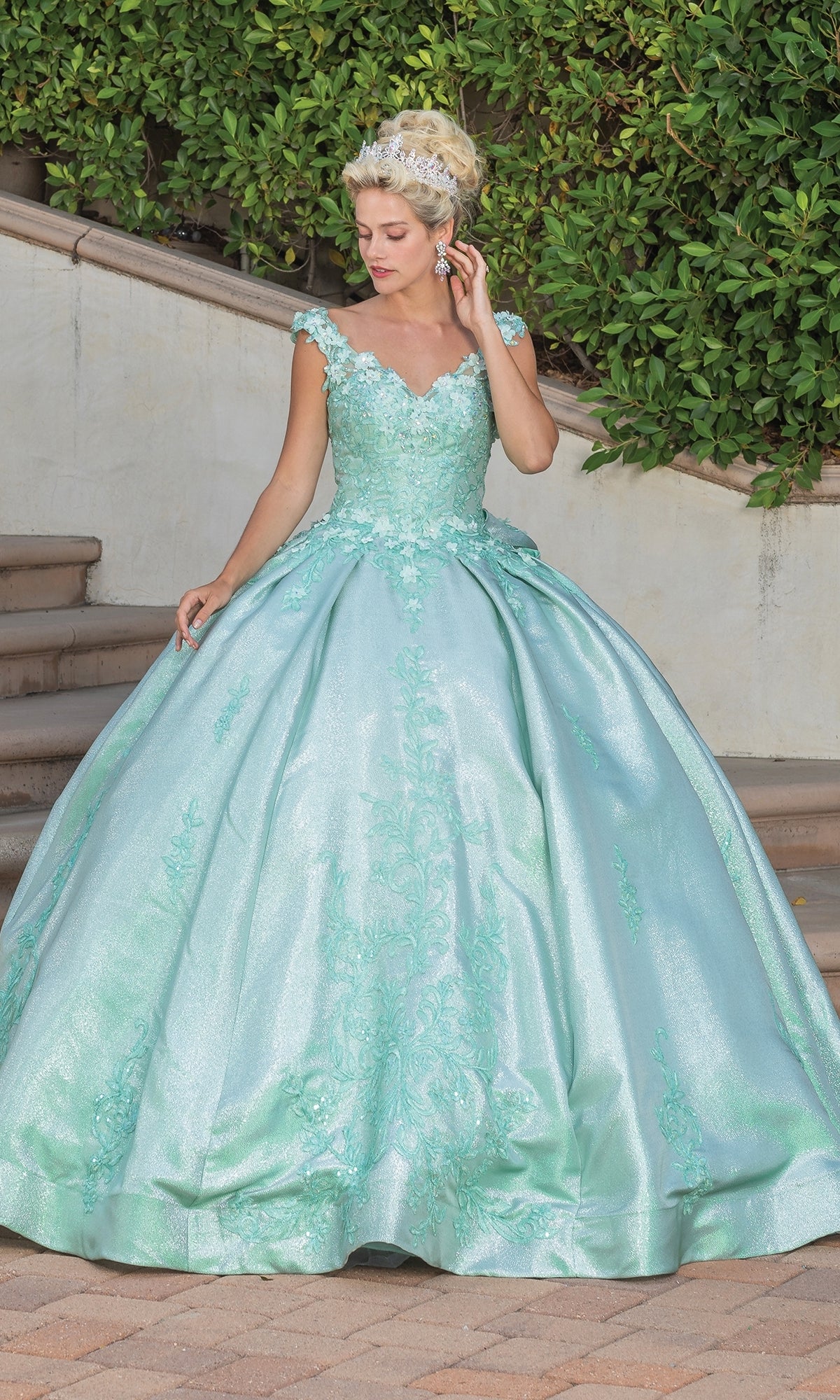 A woman in the Quinceanera Dress 1762 By Dancing Queen—a light blue ball gown with lace details and a v-neckline—wears a tiara as she stands on a paved area before stairs and green bushes.
